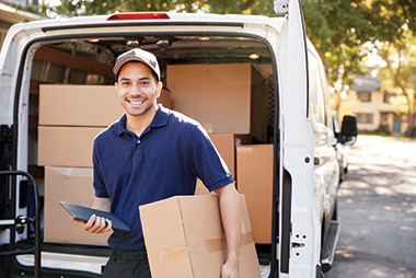 a man with a parcel under one arm and a tablet in the other land smiling behind an open van